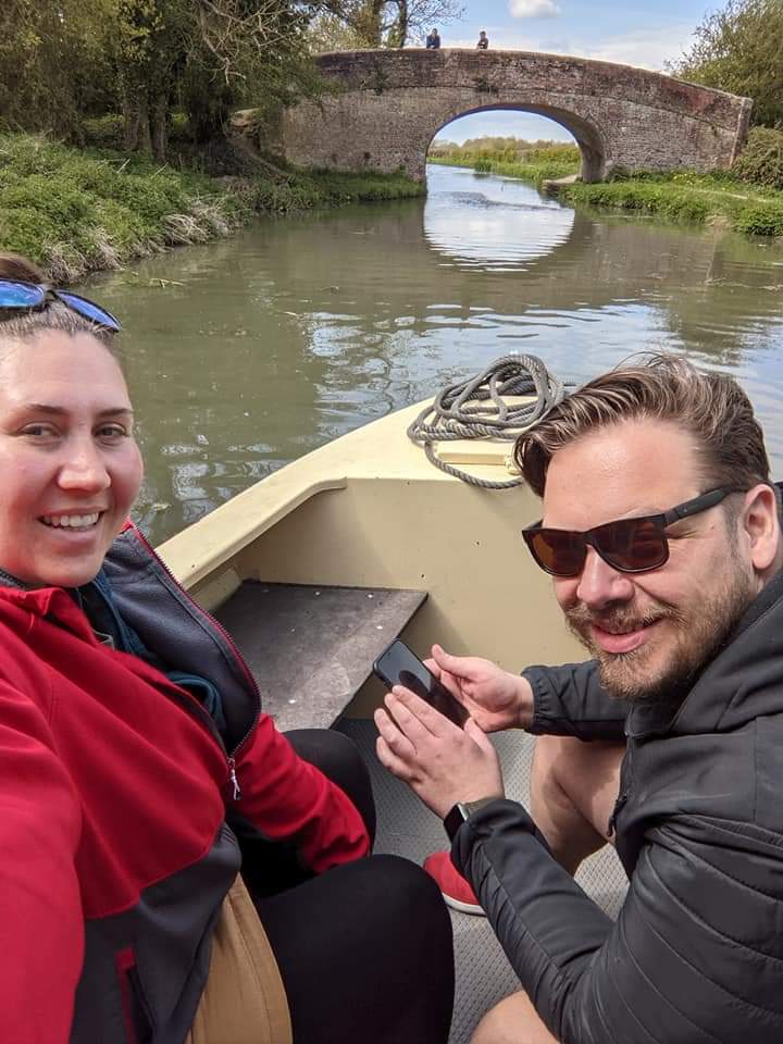 Nikyla and a male friend are on a boat on the canal. They face the camera and smile, a bridge is in the background