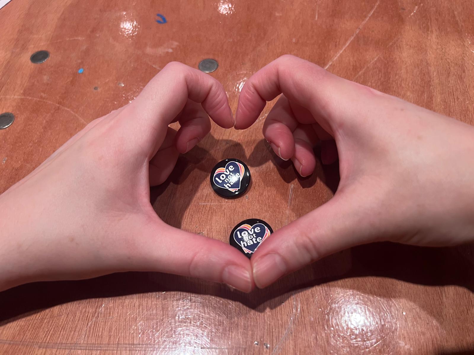 2 hands make a heart shape around 2 pin badges that say love not hate